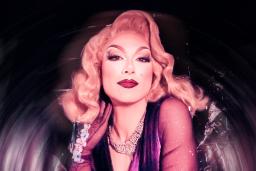 Une drag queen aux cheveux blonds mi-longs, portant des bijoux argentés et une robe violette, pose devant un fond argenté.
