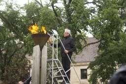 Un homme tenant un flambeau est debout sur une échelle pour allumer une flamme dans une vasque en cuivre.