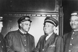 Photo en noir et blanc de quatre hommes en tenue de porteurs ferroviaires. Tous les quatre sourient et les deux du centre se serrent la main.
