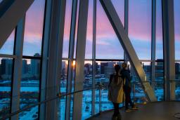 Deux personnes regardent par fenêtres allant du plancher au plafond un coucher de soleil sur un paysage urbain de Winnipeg.
