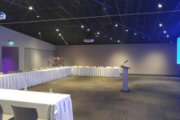 Une grande salle contenant une longue table en forme de C sur la gauche pouvant accueillir une trentaine de personnes, faisant face à un pupitre sur la droite.