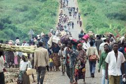 Une grande foule de personnes de tous âges portant de la nourriture et des effets personnels se dirige vers la caméra sur un long chemin de terre traversant un paysage vert vif d’herbe et de buissons. La route et la foule s’étendent dans le lointain.