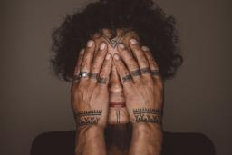 Aaju Peter se couvre les yeux et les joues avec ses mains nues. Des tatouages sont visibles sur son front, son menton, ses doigts et ses poignets. Ses mains et les parties visibles de son visage sont éclairées et encadrées par ses cheveux sombres et bouclés et ses épaules anguleuses.