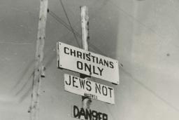 Photo noir et blanc de trois écriteaux fixés sur un poteau, où l’on peut lire en anglais « Chrétiens seulement », « Interdit aux Juifs » et « Danger ».