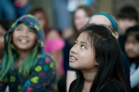 Une fille lève la tête et les yeux légèrement vers le haut. Elle a un sourire sur le visage. Derrière elle, d'autres enfants souriants sont flous.