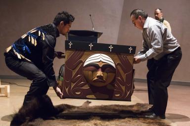 Luke Marston (à gauche) et une autre personne soulèvent la boîte en bois cintré; une peau de bison est étendue sur le plancher sous la boîte.