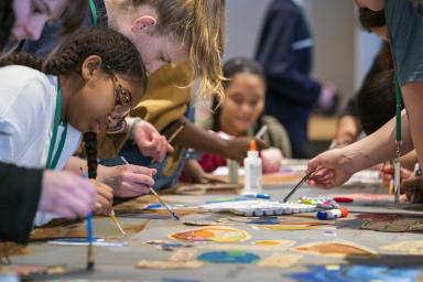 Un groupe d’enfants et de jeunes peint avec de petits pinceaux, de part et d’autre d'une longue table. Au premier plan, il y a des peintures de différentes couleurs.