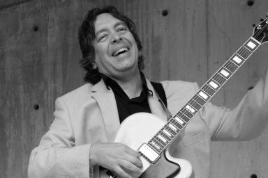 Photo en noir et blanc d’un musicien debout, riant et accordant sa guitare.