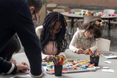 Deux enfants sont assis à une table et font de l’art tandis qu'un adulte s'appuie sur la table pour les observer.