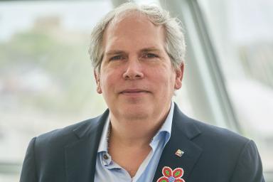 Un homme se tient face à l’objectif avec une expression sérieuse. Il porte une veste de costume sombre et une chemise boutonnée bleu clair. Il arbore une fleur multicolore de style métis sur son revers.