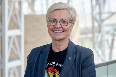 Une femme aux cheveux clairs et aux lunettes noires sourit vers l’objectif. Elle porte une veste de tailleur sombre et un t-shirt sur lequel figure une carte du Canada aux couleurs du drapeau de la Fierté inclusif.