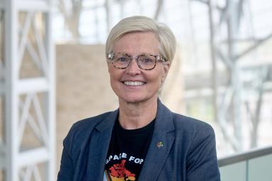 Une femme aux cheveux clairs et aux lunettes noires sourit vers l’objectif. Elle porte une veste de tailleur sombre et un t-shirt sur lequel figure une carte du Canada aux couleurs du drapeau de la Fierté inclusif.