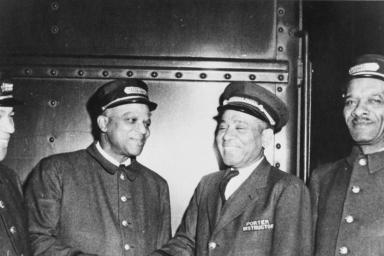 Photo en noir et blanc de quatre hommes en tenue de porteurs ferroviaires. Tous les quatre sourient et les deux du centre se serrent la main.