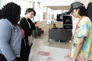 Un élève et deux adultes se tiennent de chaque côté d’une table sur laquelle des affiches sont posées.