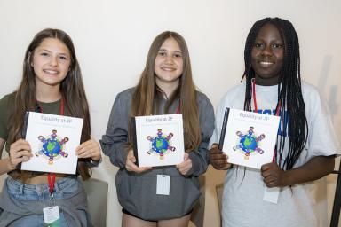 Trois élèves tiennent des exemplaires d’un livre qu’elles ont créé. La couverture du livre porte un titre anglais qui dit « L’égalité à JP » et présente un dessin de la terre avec des élèves tout autour.