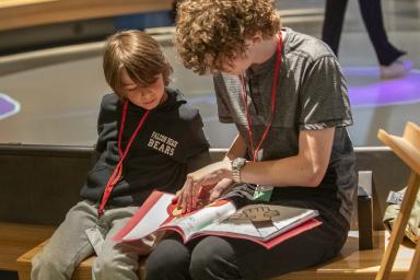 Un élève plus âgé et un élève plus jeune sont assis l’un à côté de l’autre sur un banc. L’élève le plus âgé lit un livre au plus jeune. 