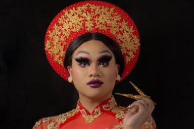 Une artiste de drag porte des vêtements culturels vietnamiens avec une coiffe et une robe longue rouge et or assorties.