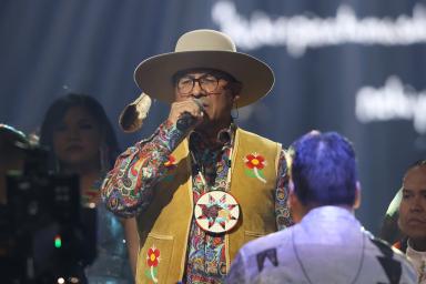 Gros plan sur un groupe de personnes portant des vestes et des gilets ornés de perles qui se produisent sur scène. Au centre, un homme portant des lunettes, un chapeau et un grand médaillon perlé chante dans un microphone.