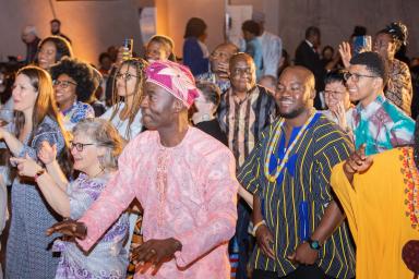 Un groupe de personnes de tous âges, dont beaucoup portent des vêtements africains traditionnels aux couleurs vives, dansent et sourient.