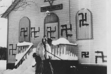 Deux personnes jouent au hockey devant une synagogue où plusieurs croix gammées ont été peintes, Sainte-Marguerite, Québec, 1938.