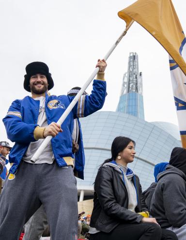 Un homme souriant dans une foule, tenant un grand drapeau jaune avec un « W » bleu et blanc au centre. L’homme porte une veste bleue des Blue Bombers de Winnipeg et se tient devant le Musée canadien pour les droits de la personne. Visibilité masquée.