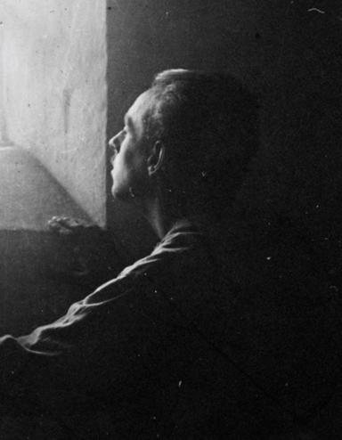 Un homme se tient dans l’ombre d’une cellule de pierre austère, regardant vers le haut la lumière vive du soleil qui entre par une fenêtre à barreaux. Visibilité masquée.
