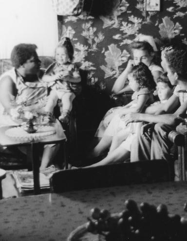 Une image en noir et blanc de femmes et de jeunes enfants assis sur des sofas et des chaises dans un petit salon. Visibilité masquée.