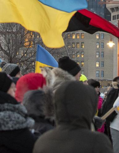 Une foule de personnes emmitouflées se tient debout dans une rue de ville pendant l’hiver, tenant de multiples drapeaux ukrainiens bleus et jaunes ainsi qu’un drapeau canadien. Le rassemblement semble être une manifestation en appui à l’Ukraine. De grands édifices à bureaux et des feux de circulation sont à l’arrière-plan sous un ciel couvert. Visibilité masquée.