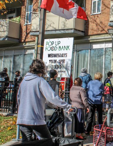 Une file de personnes attend à l’extérieur d’un bâtiment. Des panneaux indiquent qu’il s’agit d’une banque alimentaire. Un drapeau canadien flotte au-dessus d’elles. Visibilité masquée.