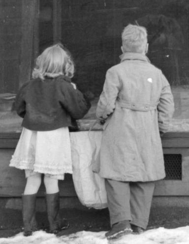 Photographie en noir et blanc de deux enfants regardant la vitrine d’un magasin abandonné. Visibilité masquée.