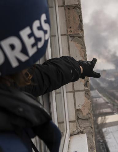Une personne portant un casque bleu foncé marqué du mot « PRESS » montre, depuis la fenêtre d’un balcon surplombant une route, une colonne de fumée à la suite d’une explosion. Visibilité masquée.