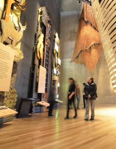 Cinq personnes regardent des expositions imposantes dans la galerie « Perspectives autochtones » du Musée. La plus proche est en bois et représente des arbres, des animaux et une plaque. Derrière les personnes, on aperçoit un théâtre de forme circulaire, construit avec des lattes de bois courbées. Visibilité masquée.