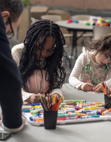 Deux enfants sont assis à une table et font de l’art tandis qu'un adulte s'appuie sur la table pour les observer. Visibilité masquée.