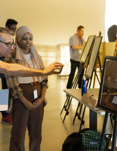 Des élèves exposent leur travail sur des chevalets dans une galerie de musée. L’accent est mis sur un élève qui discute avec un adulte souriant tout en prenant une photo de son travail avec son téléphone. Visibilité masquée.