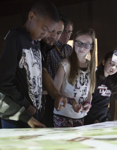 Un guide du Musée montrant à cinq élèves comment utiliser une table numérique. Ils sont tous et toutes debout autour de la table. Visibilité masquée.