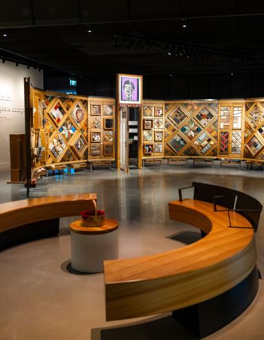 Exposition muséale présentant un grand mur de bois incurvé et autoportant sur lequel sont fixés de nombreux petits objets disposés en losanges et en rectangles. Au premier plan, des bancs circulaires créent un espace accueillant où les gens peuvent regarder et contempler l’exposition. Visibilité masquée.