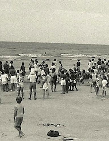 Une photographie en noir et blanc d’une foule de personnes, la plupart debout, sur une plage. Visibilité masquée.