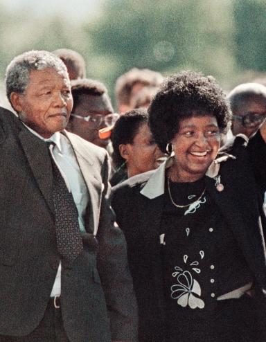 Nelson Mandela, un homme noir âgé aux cheveux grisonnants, forme un poing levé avec sa main droite et le tient au-dessus de sa tête. De sa main gauche, il tient la main de Winnie Mandela, une femme noire aux cheveux noirs. Elle sourit et forme un poing levé avec sa main gauche, à l'image du poing levé de Nelson Mandela. Une foule nombreuse est rassemblée derrière eux. Visibilité masquée.