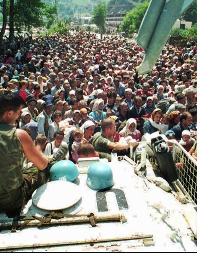Deux soldats en uniforme sont assis sur un grand véhicule, surplombant une foule dense de personnes qui s’étend au loin. Visibilité masquée.
