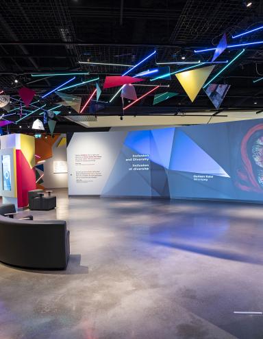 Une galerie de musée où on voit des oeuvres d'art projetées sur de grands écrans et formes géométriques colorées. Visibilité masquée.