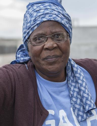 A head-and-shoulders portrait of Gogo Gladys Tyophol. She is wearing glasses and a blue patterned kerchief wrapped around her head. Her shirt says GAPA and a red AIDS ribbon is attached to her sweater. Visibilité masquée.