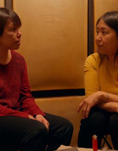 Deux femmes inuites sont assises dans une pièce sombre, l’une en face de l’autre, et discutent de manière intime. L'une porte un chandail rouge, l'autre un chandail jaune. Visibilité masquée.