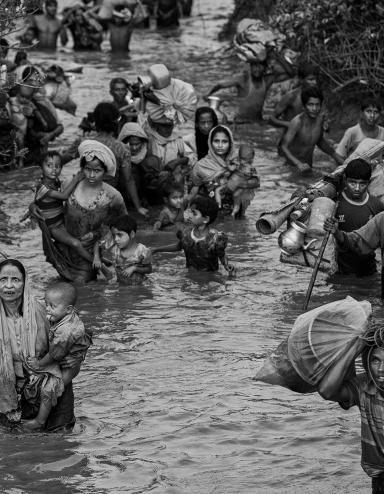 Des femmes, des enfants et des hommes rohingyas marchent dans l’eau d’une rivière qui leur monte jusqu’à la taille. Certaines personnes portent des jeunes enfants alors que d’autres portent des sacs de possessions, dont des articles ménagers. Visibilité masquée.