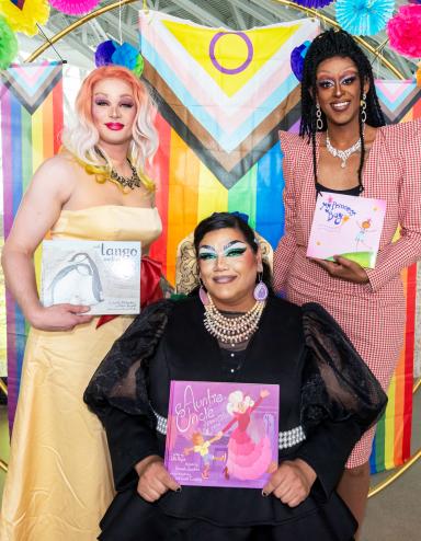 Trois artistes de drag tenant des livres se tiennent devant une arche en forme de cercle aux couleurs de l’arc-en-ciel. Visibilité masquée.
