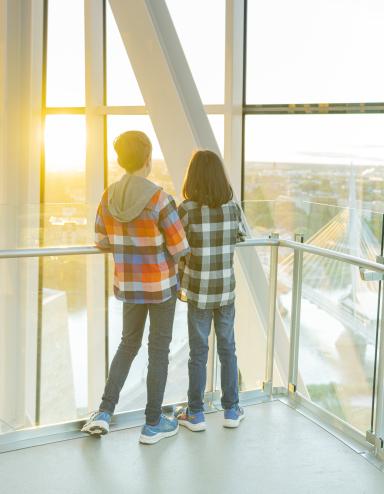 Deux enfants se tiennent dans une tour, dos à la caméra, et regardent vers l’horizon qui surplombe un paysage urbain. Visibilité masquée.