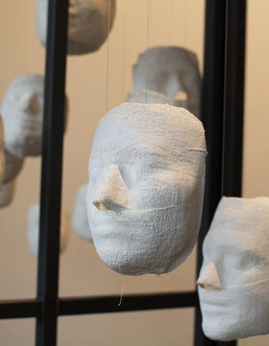 Plusieurs visages en plâtre blanc sont suspendus à des cordes, à l’intérieur d’une cage. Visibilité masquée.