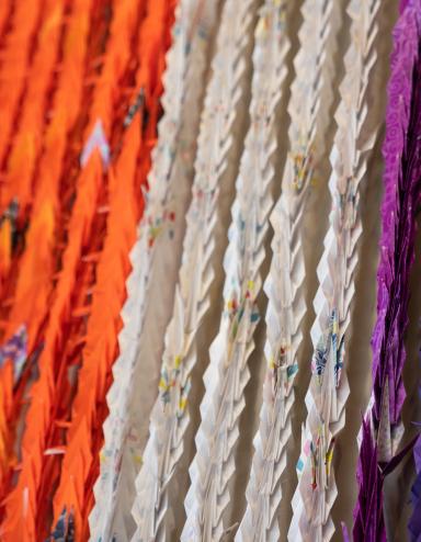 Grues multicolores en origami pressées les unes contre les autres verticalement et suspendues au mur. Visibilité masquée.