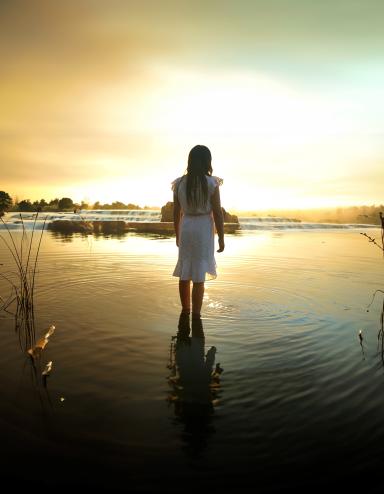 Une jeune fille aux longs cheveux noirs, vêtue d'une robe blanche jusqu'aux genoux, regarde au loin, debout jusqu'aux chevilles dans un lac calme, le soleil couchant jetant une chaude lueur jaune sur l'eau. Visibilité masquée.