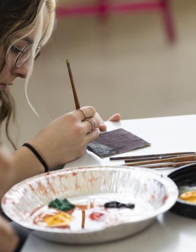 A young woman sitting at a table paints a design on a tile while a tin tray with various paints sits in front of her. An open bottle of red paint sits on the table. Visibilité masquée.