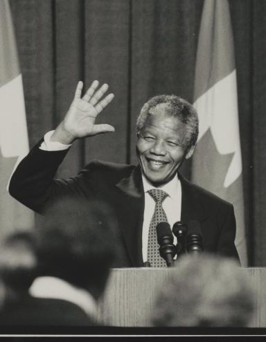 Une photographie en noir et blanc de Nelson Mandela debout derrière un podium. Il sourit et fait signe de la main. Visibilité masquée.